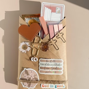 May include: A brown paper-wrapped package decorated with a tan heart, gold cross-shaped paper clips, a daisy sticker, a floral sticker, and a wax seal. It features stickers with the text be still and know that I am God psalm 46:10 and God is good.