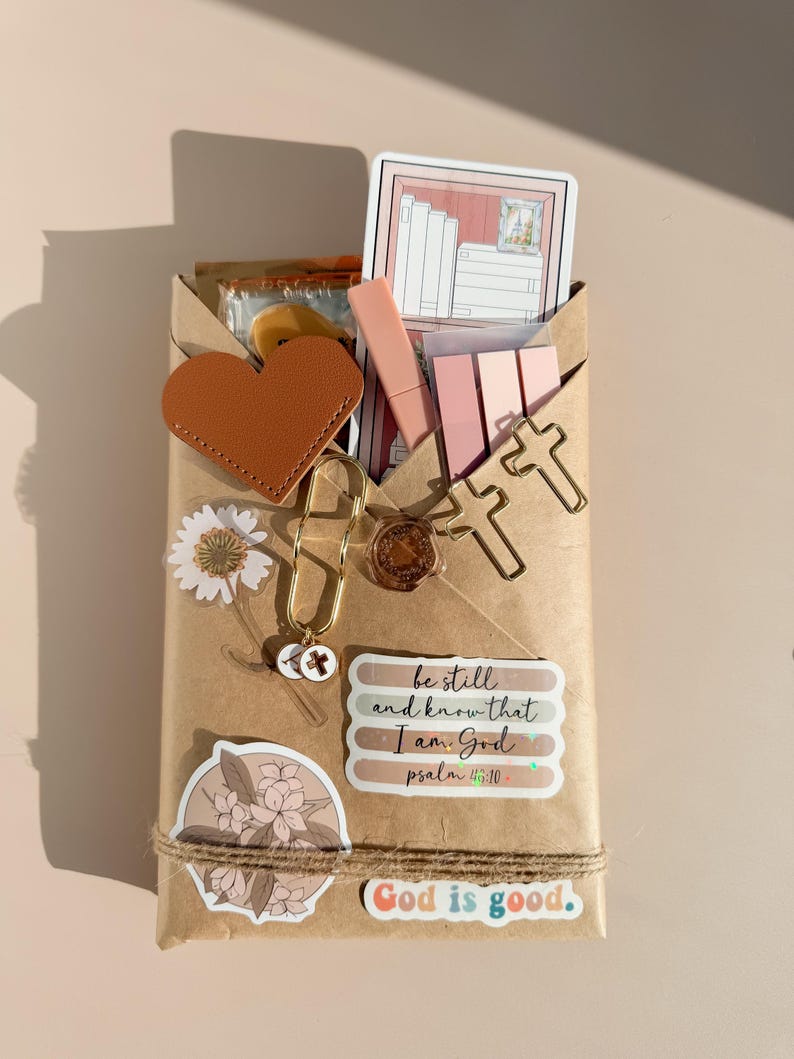 May include: A brown paper-wrapped package decorated with a tan heart, gold cross-shaped paper clips, a daisy sticker, a floral sticker, and a wax seal. It features stickers with the text be still and know that I am God psalm 46:10 and God is good.