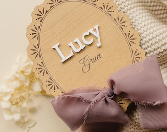 May include: An oval wooden plaque with a scalloped edge featuring the name Lucy in raised white letters and the name Grace in black script. A dusty rose-colored frayed ribbon is tied around the bottom of the light brown wood sign.