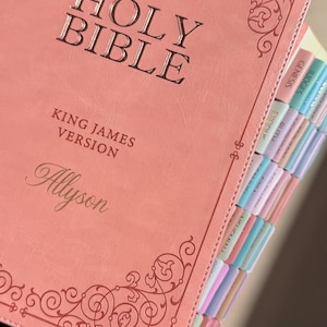 May include: A coral-colored Holy Bible King James Version with the name Allyson embossed on the front cover. The cover features decorative scrollwork at the top and bottom corners. The side of the book has multi-colored index tabs for books.