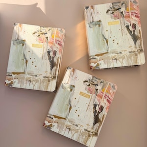 May include: Three cream-colored Holy Bible covers with abstract art in pink, brown, and gold. The covers have a gold rectangular label that reads "Holy Bible". The covers are arranged on a light brown surface.