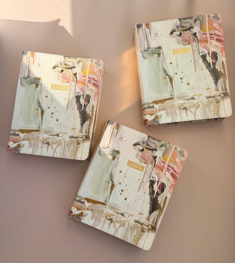 May include: Three cream-colored Holy Bible covers with abstract art in pink, brown, and gold. The covers have a gold rectangular label that reads "Holy Bible". The covers are arranged on a light brown surface.