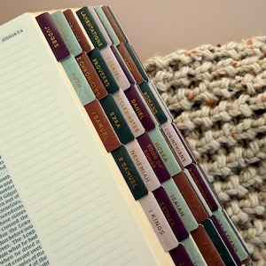 May include: Open Bible with colorful tabs labeled with book names. The tabs are in shades of green, brown, and purple with gold lettering. The pages are lined and the text of Joshua 5:4 is visible.