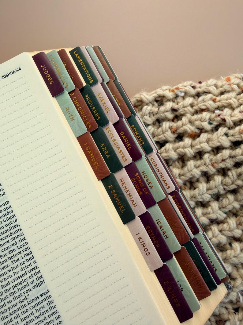 May include: Open Bible with colorful tabs labeled with book names. The tabs are in shades of green, brown, and purple with gold lettering. The pages are lined and the text of Joshua 5:4 is visible.