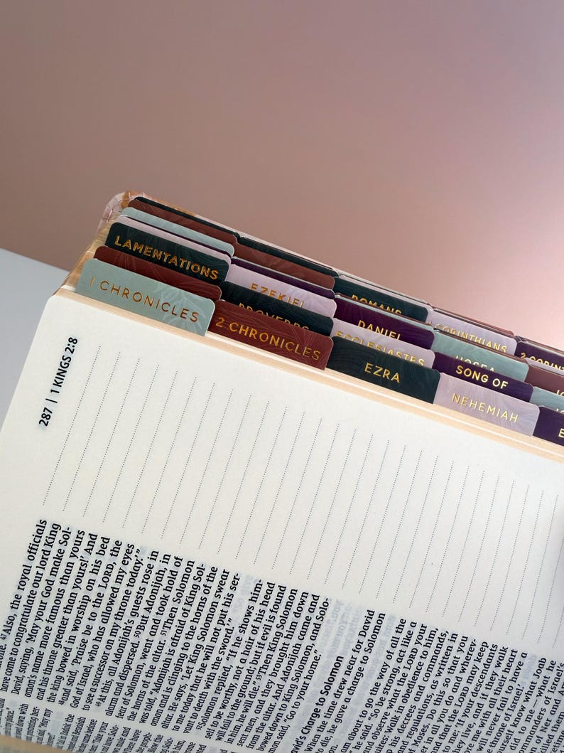 May include: Open Bible with labeled tabs for different books, including Lamentations, Chronicles, and Ezekiel. The tabs are in various colors with gold lettering. The text on the page is visible, with the reference 1 Kings 2:8.