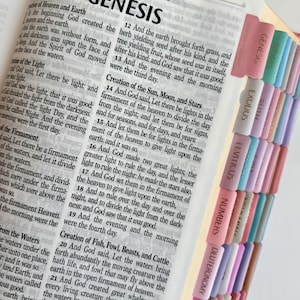 May include: An open Bible displaying the text of Genesis with black printed words on white paper. The page edges feature a series of colorful, vertical adhesive tabs labeled with book names including Genesis, Exodus, Leviticus, Numbers, and Joshua.