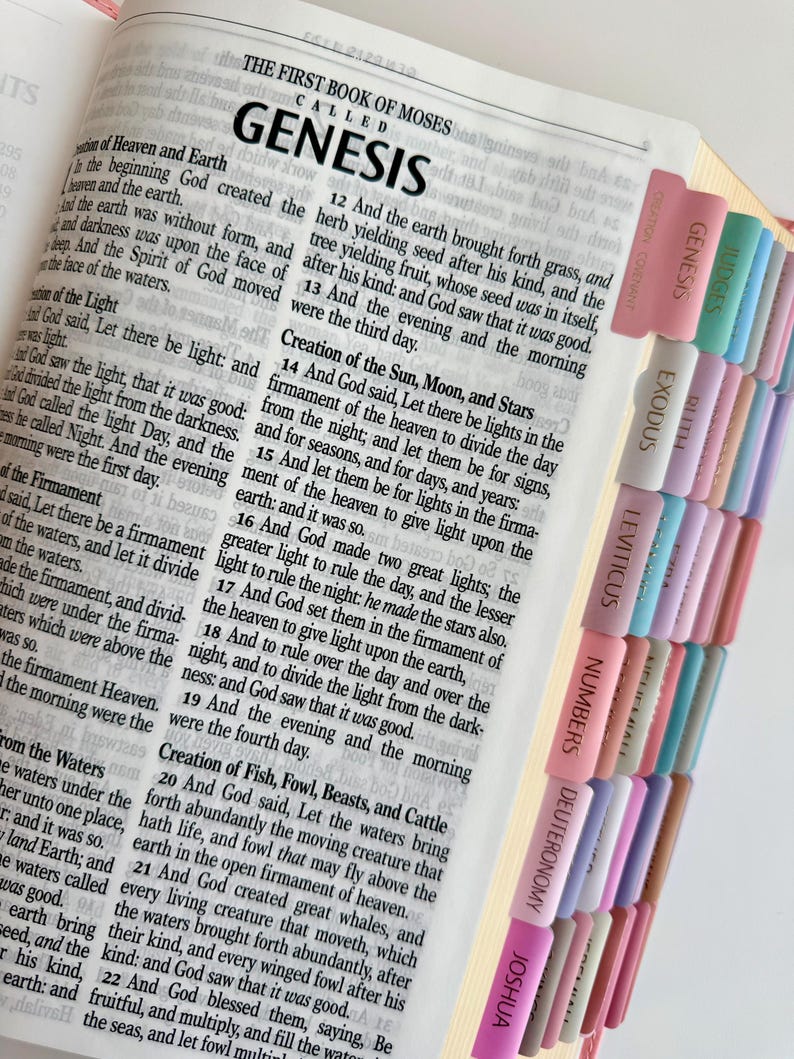 May include: An open Bible displaying the text of Genesis with black printed words on white paper. The page edges feature a series of colorful, vertical adhesive tabs labeled with book names including Genesis, Exodus, Leviticus, Numbers, and Joshua.