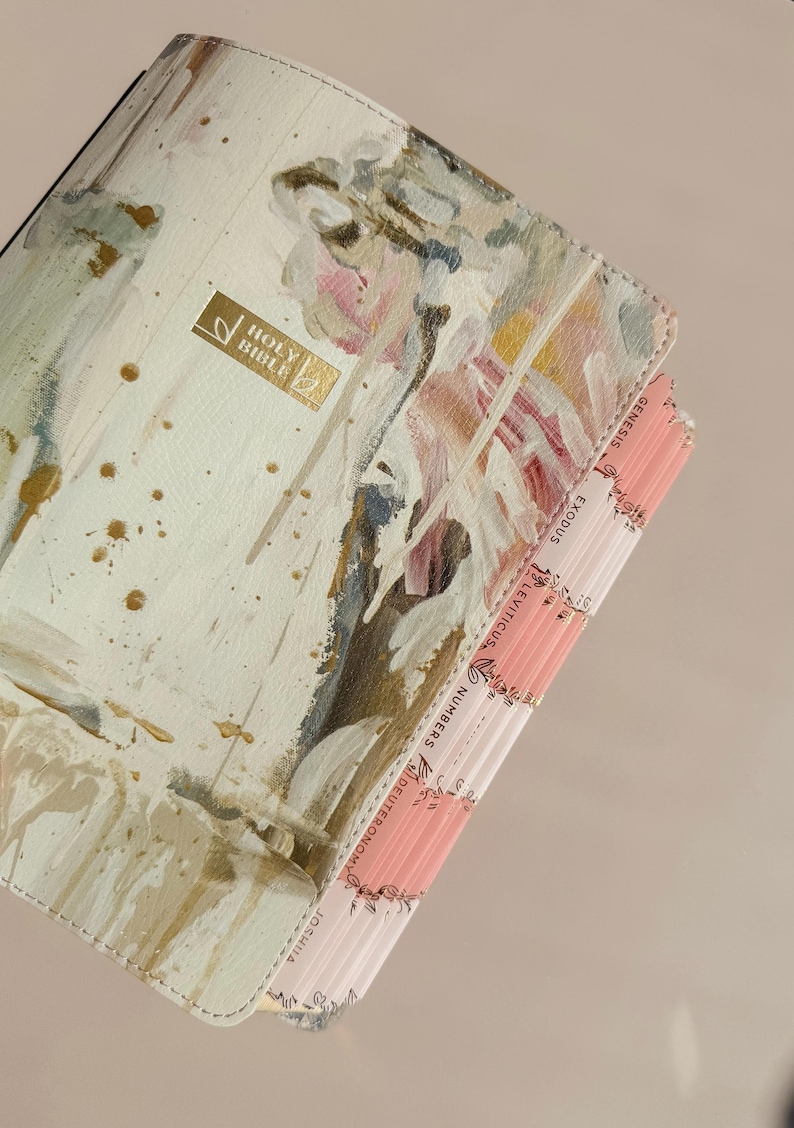 May include: A Holy Bible with a cream-colored cover featuring abstract brushstrokes in gold, white, and soft pink. The cover includes a gold rectangular plaque with the text Holy Bible. The page edges have pink tabs labeled Genesis, Exodus, Leviticus, Numbers, Deuteronomy, and Joshua.
