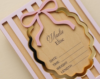 May include: A rectangular wooden plaque with light pink vertical stripes featuring a central scalloped gold mirror frame. A pink bow sits atop the frame, which is engraved with the name Maelie Elise and lines for date, time, weight, and length.