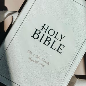 May include: A white leather-bound Holy Bible with an embossed vine pattern on the cover. The text reads HOLY BIBLE, KING JAMES VERSION, Mr. & Mrs. Townsley, and August 8th, 2026. The cover features stitched edges and a clean, light-colored finish.
