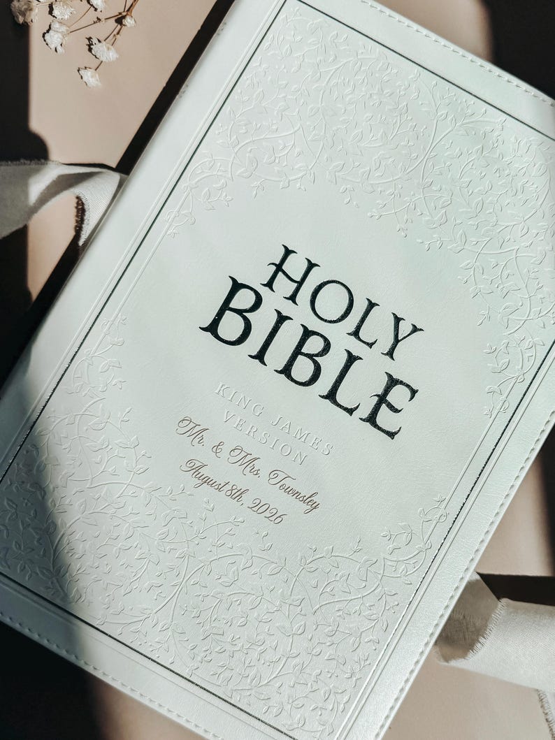 May include: A white leather-bound Holy Bible with an embossed vine pattern on the cover. The text reads HOLY BIBLE, KING JAMES VERSION, Mr. & Mrs. Townsley, and August 8th, 2026. The cover features stitched edges and a clean, light-colored finish.