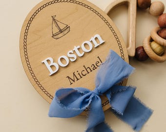 May include: A round wooden plaque featuring a laser-engraved sailboat, the name Boston in raised white letters, the name Michael engraved below, and a frayed blue ribbon tied to the front, accompanied by a wooden teething ring with beads.