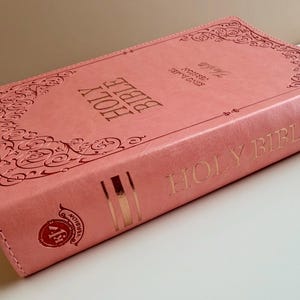 May include: A coral-colored Holy Bible with a leather-like cover featuring embossed scrollwork borders. The front cover displays the text Holy Bible and King James Version in gold foil. The spine includes the text Holy Bible and a KJV logo.