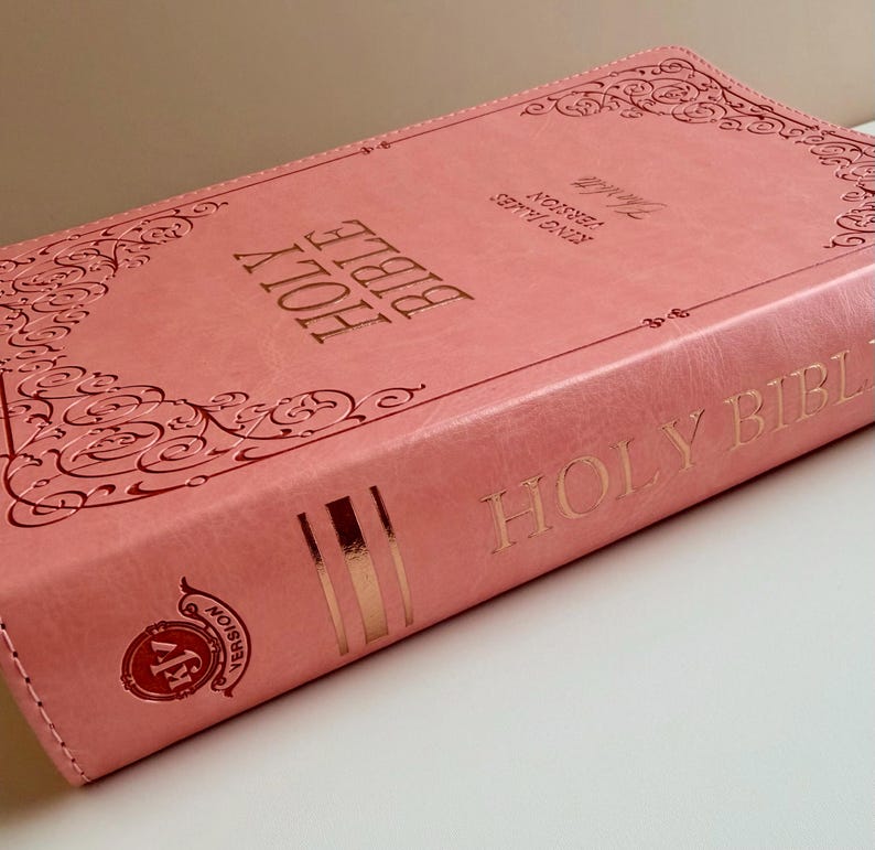May include: A coral-colored Holy Bible with a leather-like cover featuring embossed scrollwork borders. The front cover displays the text Holy Bible and King James Version in gold foil. The spine includes the text Holy Bible and a KJV logo.
