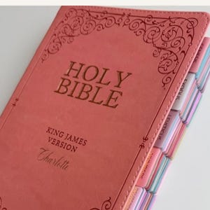 May include: A coral-colored Holy Bible, King James Version, with the name Charlotte embossed on the cover. The book features decorative scrollwork at the top and bottom, and includes multi-colored index tabs along the side for navigation.