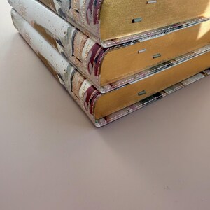 May include: Three stacked hardcover books with gold-gilded edges. The covers feature a watercolor-style design with shades of beige, brown, and burgundy. The spines are visible, and the books are stacked on top of each other.