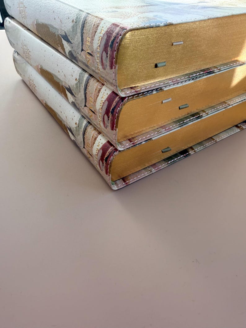 May include: Three stacked hardcover books with gold-gilded edges. The covers feature a watercolor-style design with shades of beige, brown, and burgundy. The spines are visible, and the books are stacked on top of each other.