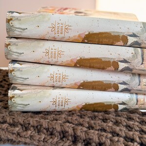 May include: A stack of Holy Bibles with decorative covers. The covers feature a watercolor design in shades of white, gold, and pink. The spine of each book is labeled "Holy Bible" in gold lettering. The books rest on a brown, textured surface.