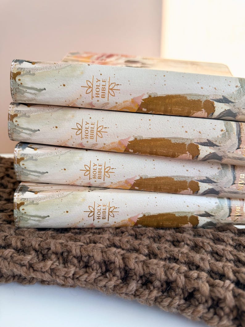May include: A stack of Holy Bibles with decorative covers. The covers feature a watercolor design in shades of white, gold, and pink. The spine of each book is labeled "Holy Bible" in gold lettering. The books rest on a brown, textured surface.