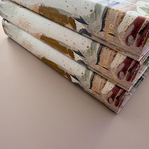 May include: Three stacked hardcover books with abstract watercolor-style covers. The covers feature a mix of colors including gold, pink, and blue. The spines have gold lettering that reads "Holy Bible" and "NIVE".