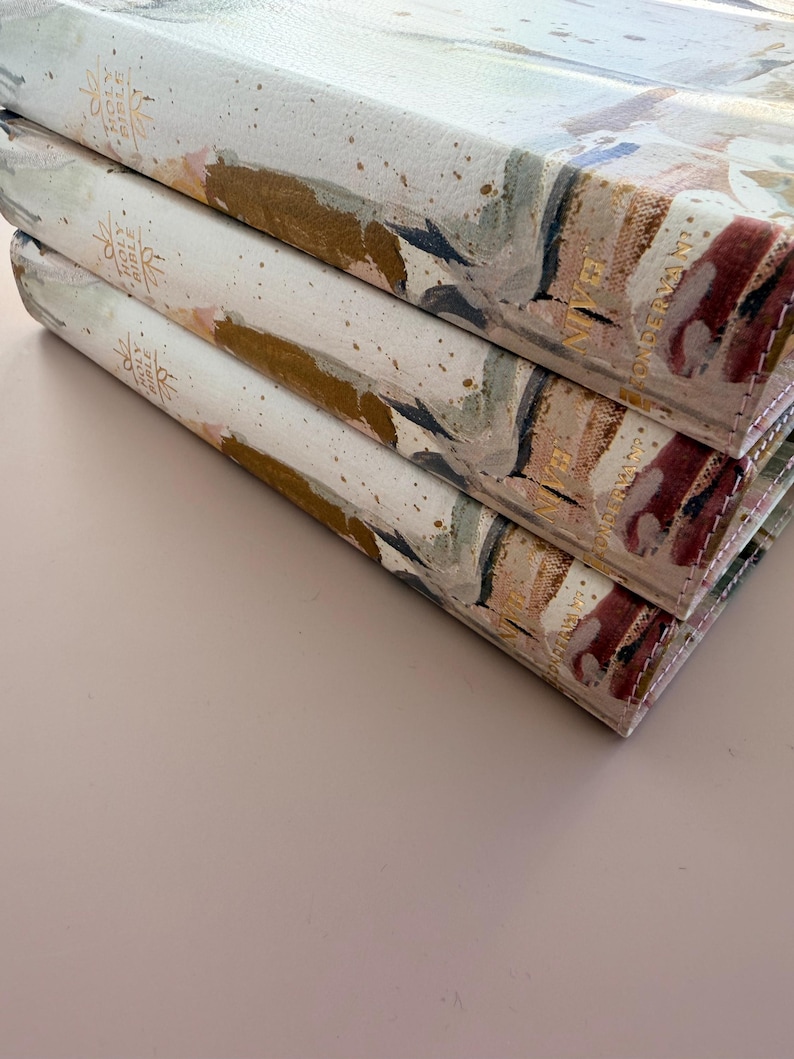 May include: Three stacked hardcover books with abstract watercolor-style covers. The covers feature a mix of colors including gold, pink, and blue. The spines have gold lettering that reads "Holy Bible" and "NIVE".