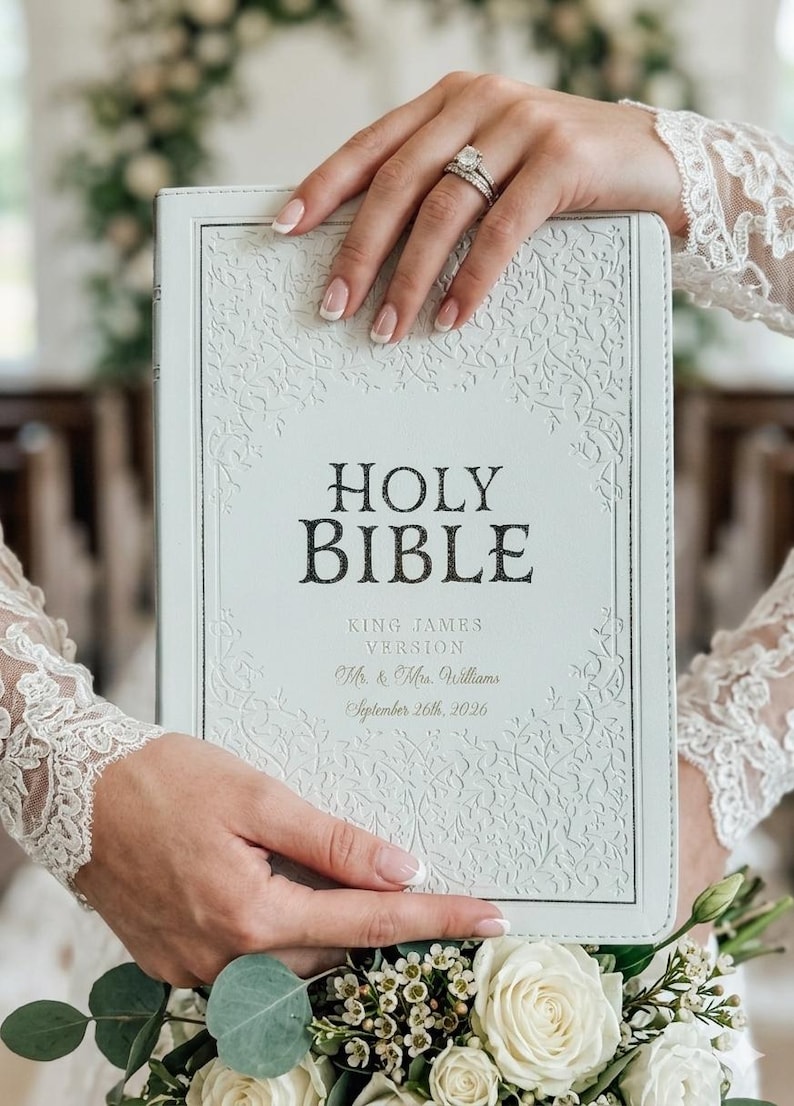 May include: A white Holy Bible with an embossed vine pattern on the cover. The text reads Holy Bible, King James Version, Mr. & Mrs. Williams, and September 26th, 2026. The cover features a decorative border and a central area for the text.