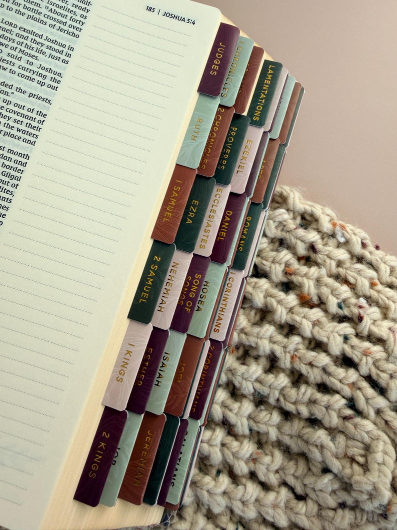 May include: Open Bible with colorful, textured tabs labeled with book names. The tabs are in shades of burgundy, green, brown, and pink. The book is open to a page with text and lines. A knitted beige blanket is in the background.