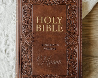 May include: A brown leather-bound Holy Bible, King James Version, featuring an embossed decorative border and the name Mason engraved on the front cover. The text Holy Bible is printed in gold lettering on the front of the book cover.