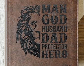 May include: A brown leather-like journal featuring a black engraved lion profile on the left and the text MAN OF GOD HUSBAND DAD PROTECTOR HERO on the right. A small cross is at the top right, and the name Collin is engraved at the bottom right.