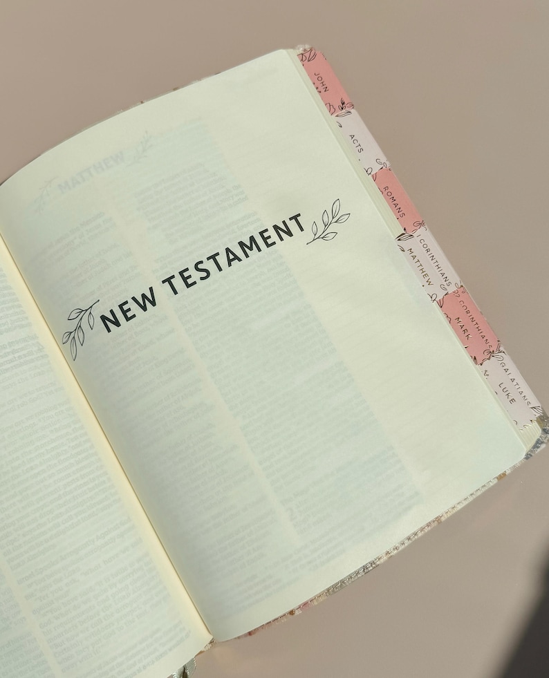 May include: An open book showing the title page for the New Testament in black text with small leaf illustrations. The page edges feature pink and white patterned tabs labeled with book names including John, Acts, Romans, and Corinthians.