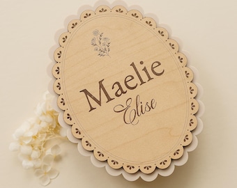 May include: An oval wooden plaque with a scalloped edge featuring the engraved name Maelie Elise and a small floral illustration. The light-colored wood has a smooth finish and is layered over a slightly larger, pale cream scalloped backing.