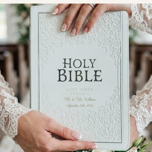 May include: A white Holy Bible, King James Version, featuring an embossed floral border and personalized text reading Mr. & Mrs. Williams, September 26th, 2026. The cover has a textured finish with dark lettering for the title and light gold text.