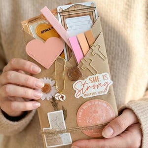 May include: A brown paper envelope filled with stationery items including a pink heart cutout, cross-shaped paper clips, a gold charm, and stickers. One sticker features the text "SHE IS Strong PROVERBS 31:25" and another shows a sun illustration.