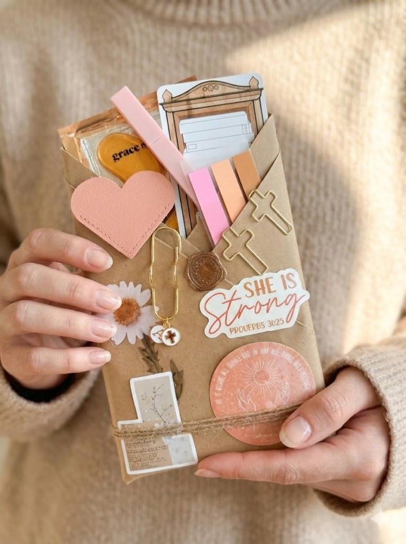 May include: A brown paper envelope filled with stationery items including a pink heart cutout, cross-shaped paper clips, a gold charm, and stickers. One sticker features the text "SHE IS Strong PROVERBS 31:25" and another shows a sun illustration.