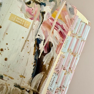 May include: A hardcover Holy Bible with a cream-colored cover featuring abstract paint strokes in pink, brown, and white. Gold lettering on the cover reads "HOLY BIBLE". The pages are edged with pastel-colored tabs labeled with book names.
