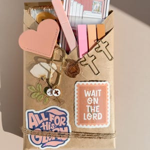 May include: A brown paper package decorated with stickers including one that reads ALL FOR HIS GLORY and another that says WAIT ON THE LORD. It features a pink heart, a gold paperclip with charms, two cross-shaped paperclips, and sticky notes.