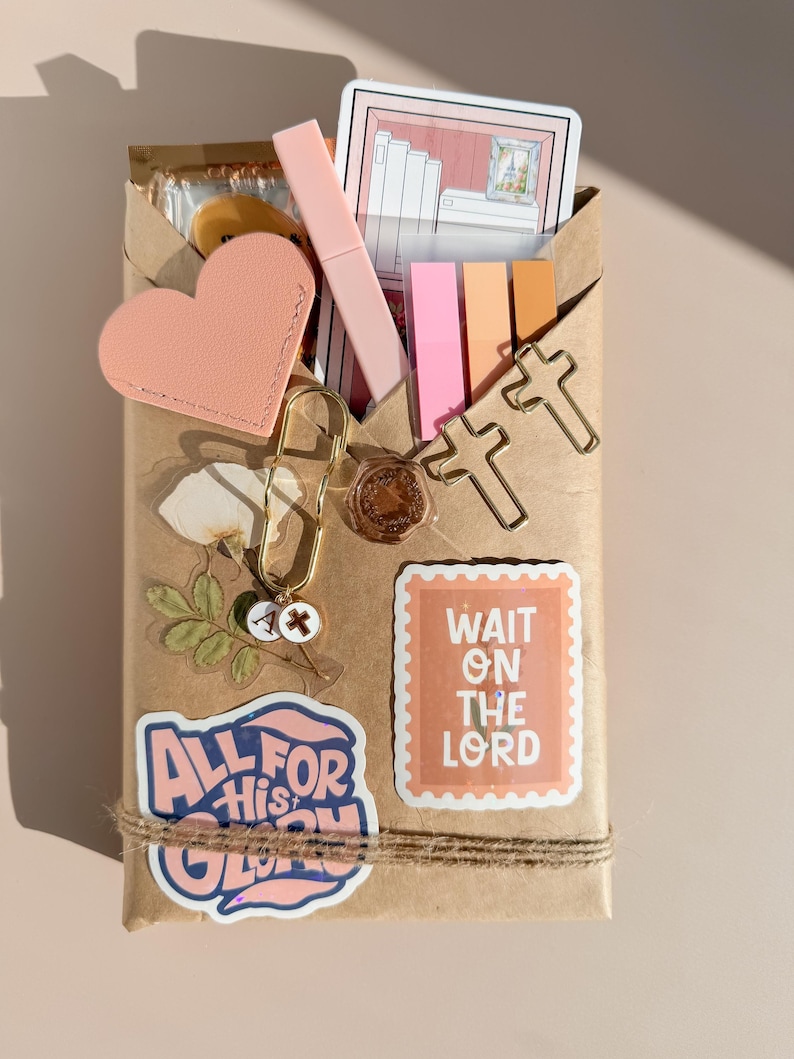 May include: A brown paper package decorated with stickers including one that reads ALL FOR HIS GLORY and another that says WAIT ON THE LORD. It features a pink heart, a gold paperclip with charms, two cross-shaped paperclips, and sticky notes.