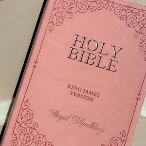 May include: A light pink Holy Bible, King James Version, featuring a dark pink decorative scrollwork border. The cover is embossed with the text HOLY BIBLE, KING JAMES VERSION, and the personalized name Abigail Mandhlazi in a script font.