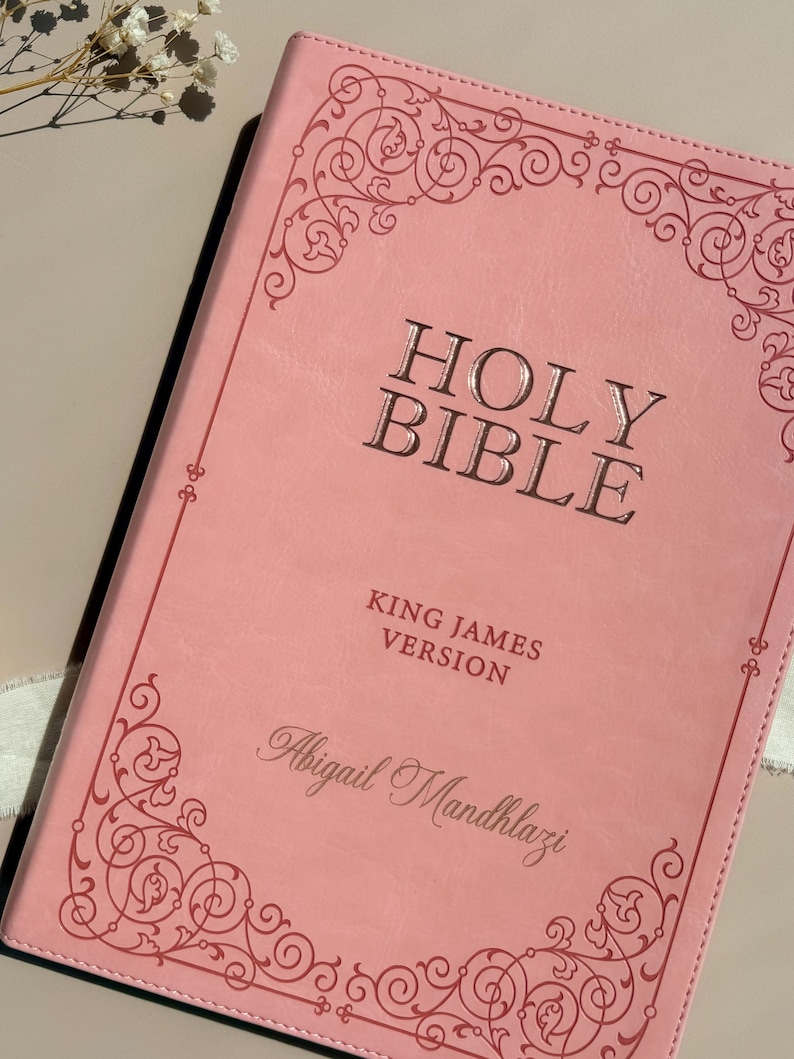 May include: A light pink Holy Bible, King James Version, featuring a dark pink decorative scrollwork border. The cover is embossed with the text HOLY BIBLE, KING JAMES VERSION, and the personalized name Abigail Mandhlazi in a script font.