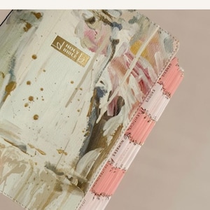 May include: A Bible with a cream-colored cover featuring an abstract painted design in shades of pink, green, and gold. The cover includes a gold-stamped Holy Bible emblem. The side features pink index tabs labeled Genesis, Exodus, Leviticus, Numbers, Deuteronomy, and Joshua.