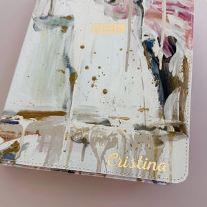 May include: A white book cover featuring an abstract paint design with streaks of gold, pink, blue, and brown. The cover includes the gold embossed text "HOLY BIBLE" in the center and the gold cursive name "Cristina" printed at the bottom right.