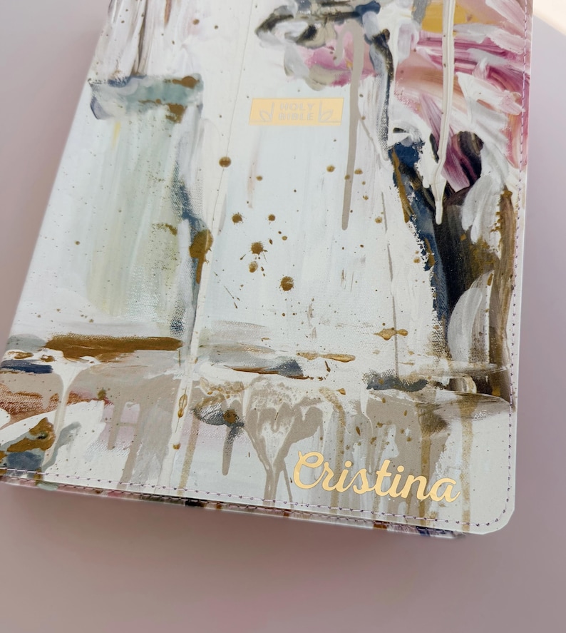 May include: A white book cover featuring an abstract paint design with streaks of gold, pink, blue, and brown. The cover includes the gold embossed text "HOLY BIBLE" in the center and the gold cursive name "Cristina" printed at the bottom right.
