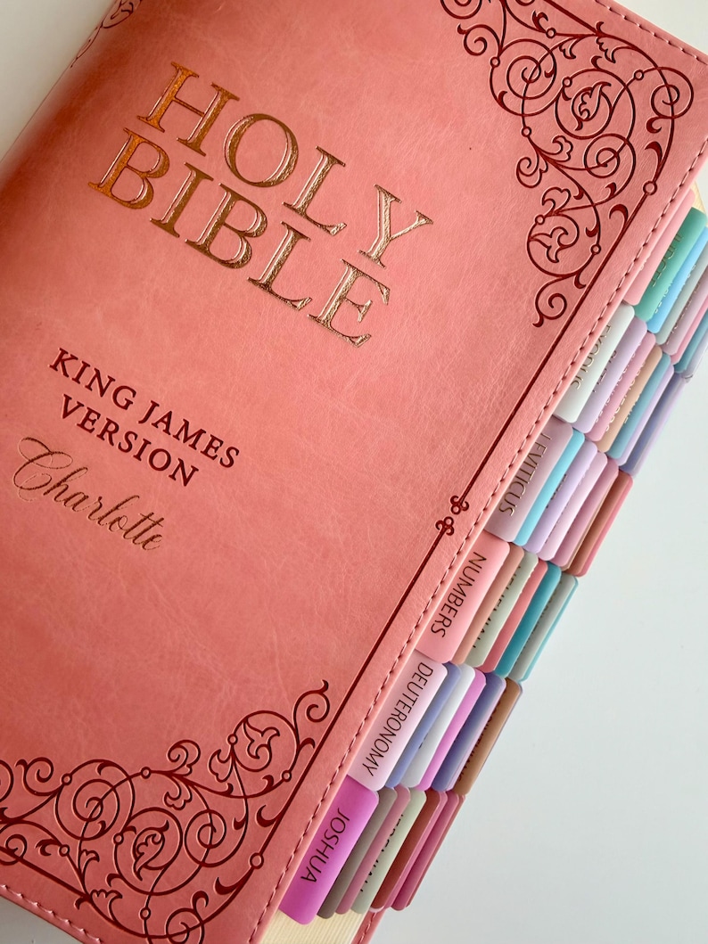 May include: A coral-colored Holy Bible King James Version with the name Charlotte embossed on the cover. The book features gold foil lettering, decorative scrollwork at the top and bottom, and a series of colorful, labeled index tabs along the edge.