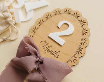 May include: An oval wooden milestone plaque featuring a scalloped edge with laser-cut sunburst details. A large white number 2 is attached to the center above the word Months in black script. A dusty rose chiffon ribbon is tied at the base.