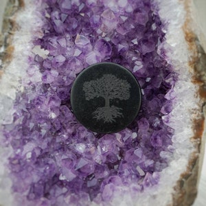 May include: A black round pendant with a tree design carved into it, resting on a bed of purple amethyst crystals.