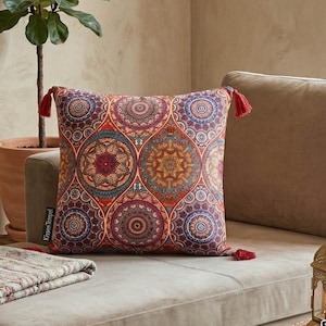 Op de afbeelding: Vierkant kussen met een kleurrijk mandala-patroon in rood, oranje, blauw en paars. Het heeft rode kwastjes op elke hoek. Het kussen ligt op een beige bank. Afmetingen: ongeveer 40 cm x 40 cm.