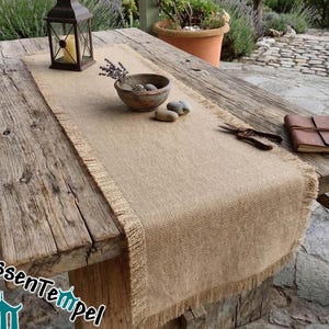 Op de afbeelding: Een rustieke tafelloper van jute met franjes, gepresenteerd op een verweerde houten tafel. De scène omvat een lantaarn, een houten kom met lavendel en een leren notitieboek, wat een natuurlijke, landelijke uitstraling creëert. De tekst "KissenTempel" is zichtbaar.