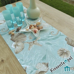 Może przedstawiać: Jasno niebieski bieżnik stołowy z wzorem muszli i rozgwiazd. Bieżnik jest ozdobiony muszlami, rozgwiazdami i wodorostami. Bieżnik leży na drewnianym stole z wazą i szklankami.