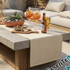 May include: An outdoor patio scene featuring a concrete and wood coffee table, two sofas, and potted plants. The table is set with drinks, snacks, and a beige table runner. The scene is illuminated by natural light.
