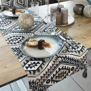 May include: A black, white, and brown geometric table runner with tassels on the ends. The runner is laid on a wooden table with a white and black patterned pillow, two clear glass candle holders, and two silver candle holders. The table runner is decorated with two silver candle holders, two clear glass candle holders, and a white and black patterned pillow.